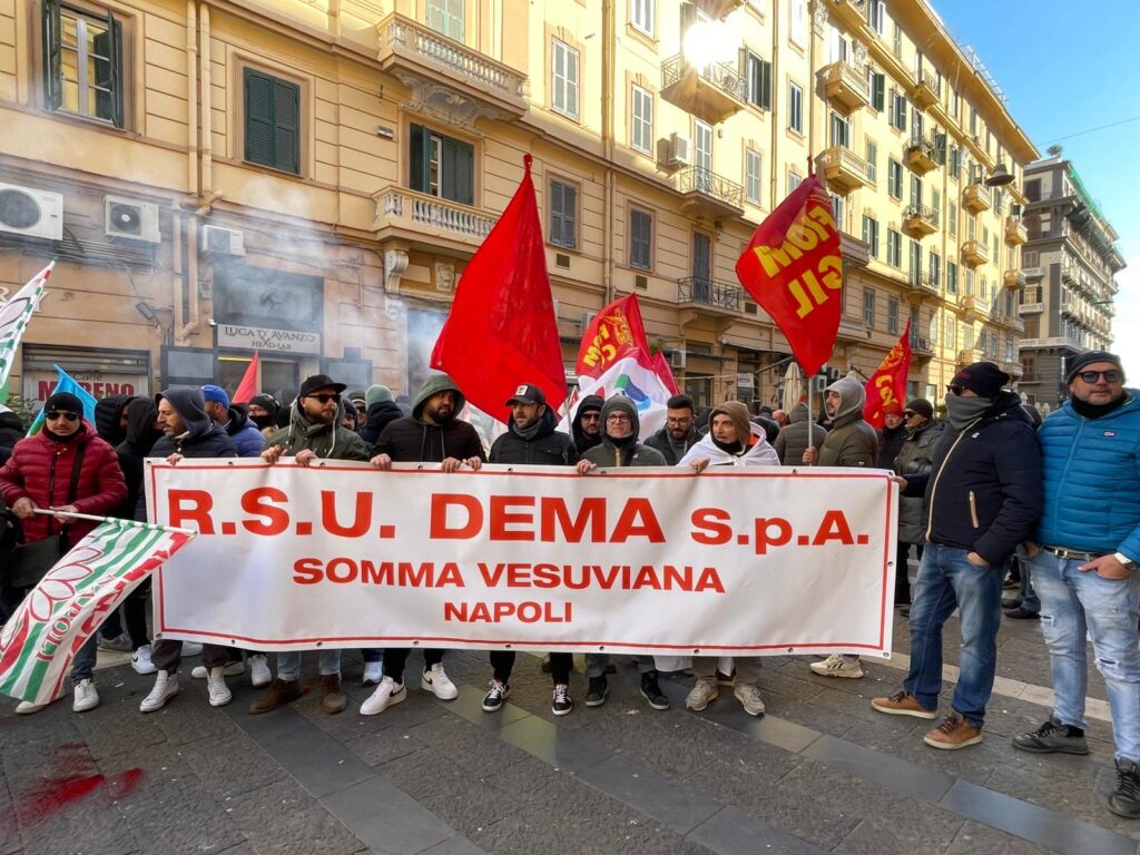 la foto mostra un gruppo di lavoratori della DEMA con bandiere e striscione