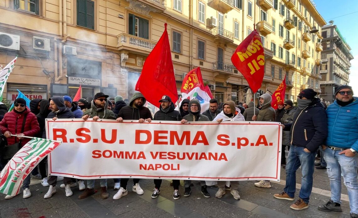 la foto mostra un gruppo di lavoratori della DEMA con bandiere e striscione